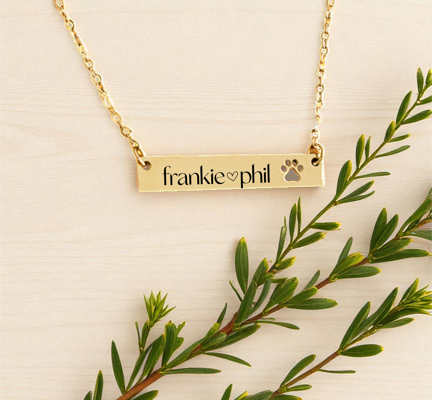 Gold necklace with engraved name and paw print on a beige background with green leaves.